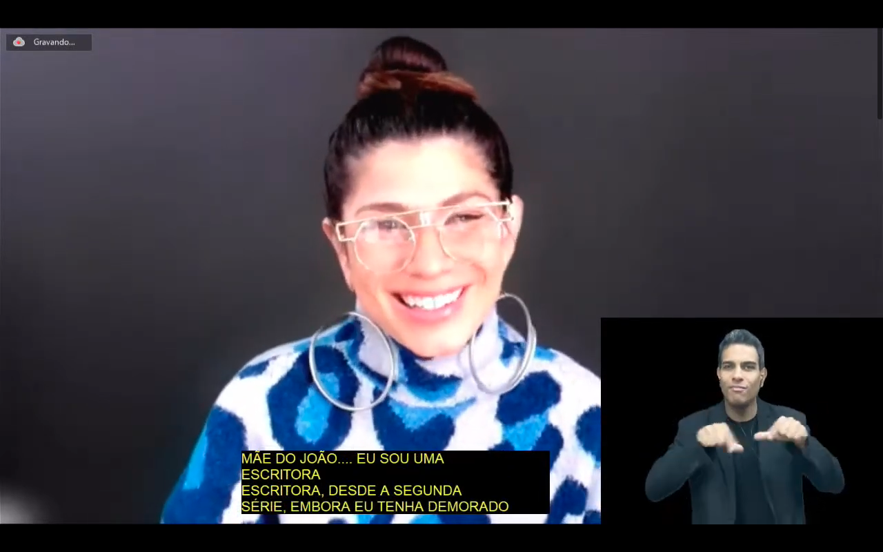 Lau Patron é branca, usa óculos de aros redondos e grande, tem cabelo escuro presos em um coque. usa um blusa branca com desenhos azuis. 