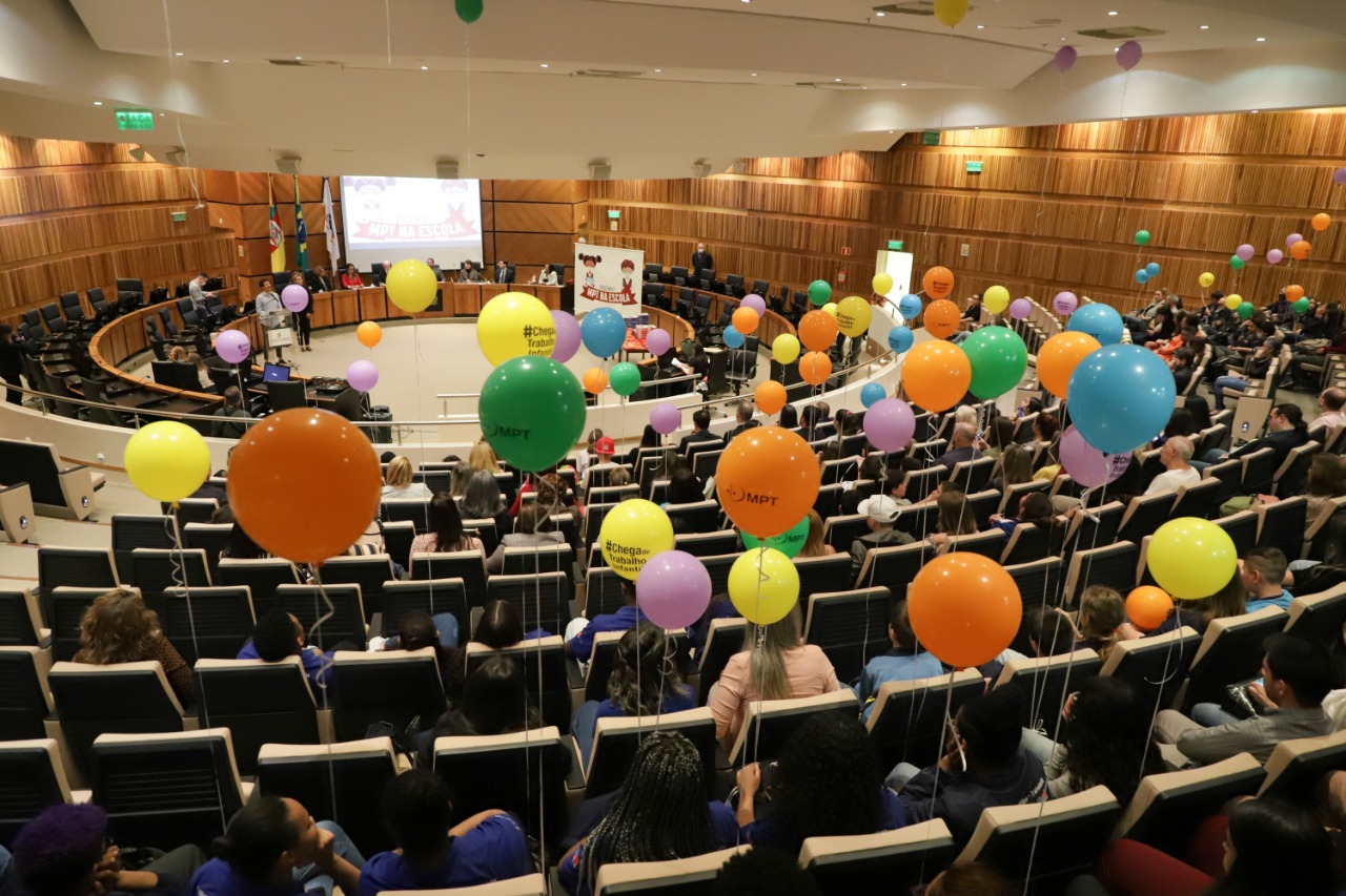 Foto do plenário o público segurando balões e, ao fundo, a mesa do evento. 