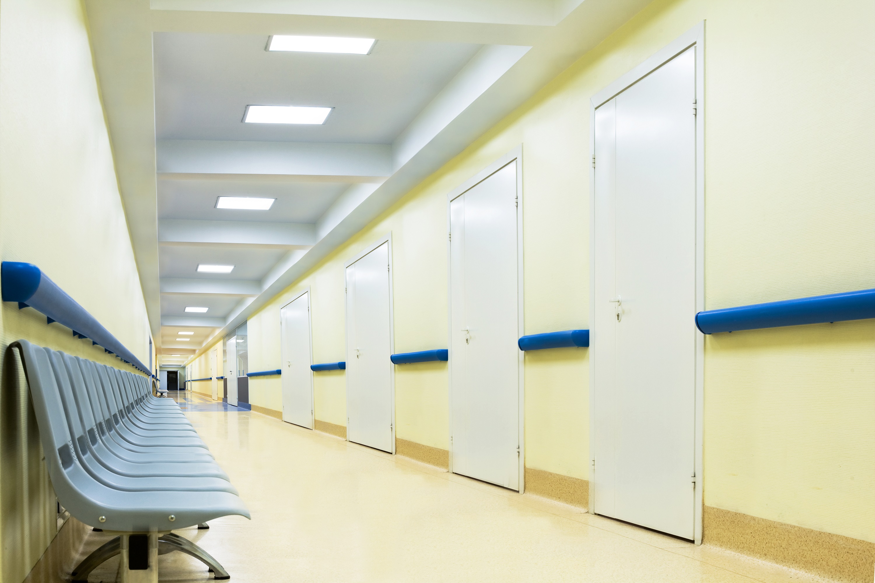 Corredor de Hospital vazio. Paredes e piso são bege e há cadeiras azuis. Há uma listra contínua no meio da parede, na cor azul. Em frente às cadeiras, há portas brancas.