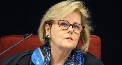Rosa Weber é branca, loira, usa óculos de aro preto, toga preta e um lenço azul no pescoço.