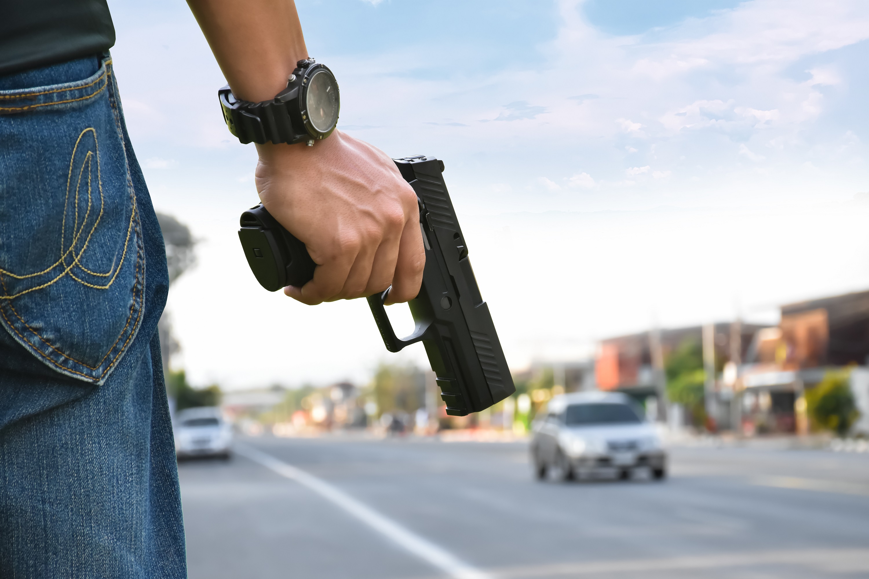 Foto mostra mão de homem, armado, à beira de uma estrada. Ele está de costas, usa calça jeans, relógio de pulso e porta uma arma na cor preta.