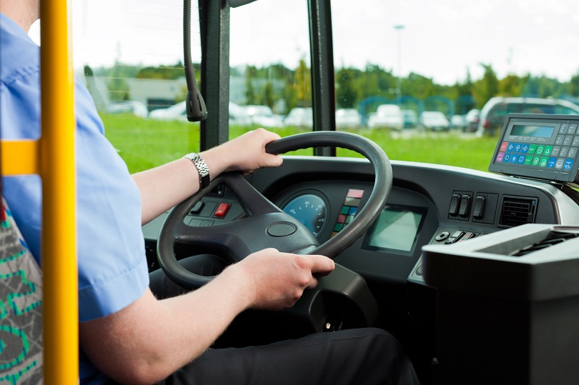 Foto ilustrativa em plano médio de um homem que está sentado ao volante, dirigindo transporte coletivo de passageiros. 