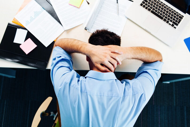 Foto ilustrativa de homem com a cabeça baixa sobre uma mesa de trabalho desorganizada.