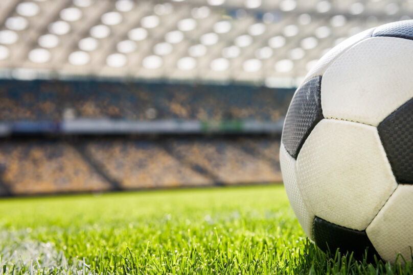 Bola de futebol, nas cores preta e branca, sobre um gramado. Ao fundo, em imagem desfocada, há a arquibancada. 