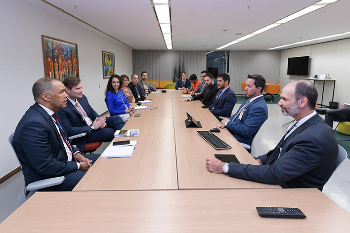 Foto em ângulo aberto, mostrando participantes sentados à mesa de reuniões