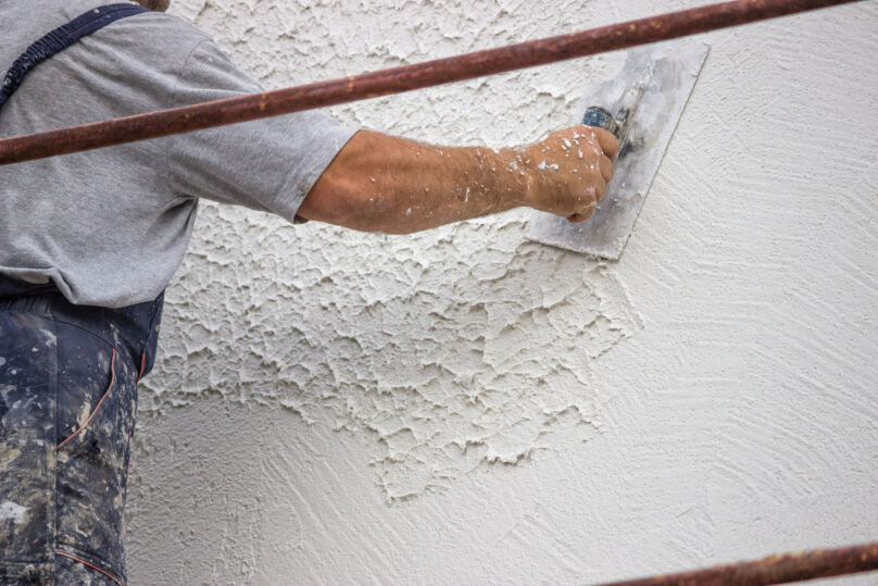 Imagem mostra homem aplicando gesso em uma parede. Ele veste macacão azul e camiseta cinza. A parede é branca.