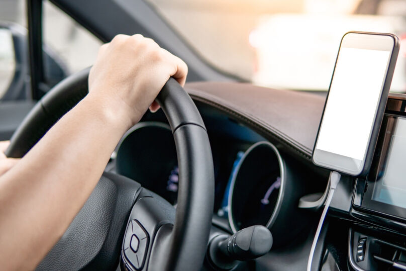 Foto ilustrativa mostra as mão de uma pessoa no volante de um carro. Ao lado direito, um celular está acoplado no painel com a tela em branco.