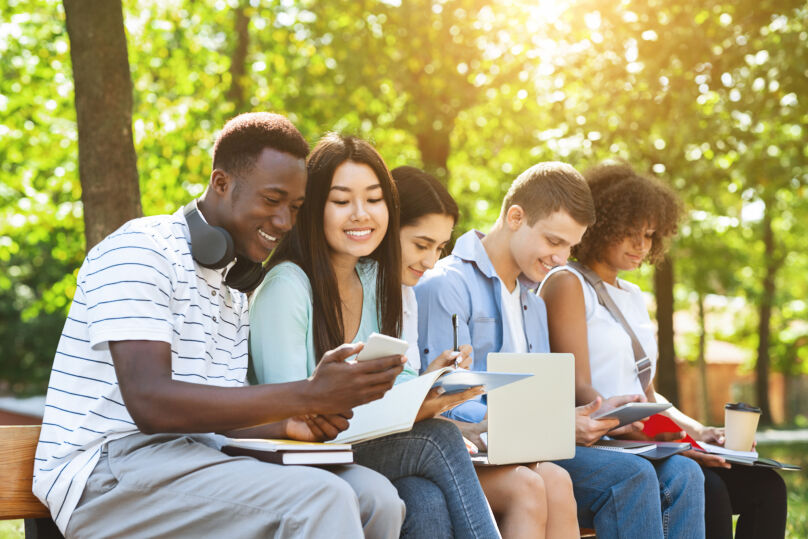 Foto ilustrativa de vários jovens sentados em um banco, ao ar livre. Eles sorriem e estão vendo conteúdos em celulares, tablets e livros.