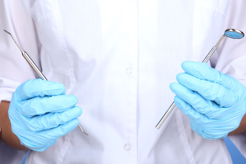 Foto ilustrativa das mãos de um dentista de luvas segurando ferramentas típicas de trabalho
