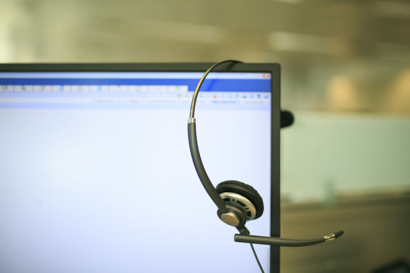Foto ilustrativa mostra um headset pendurado na tela de um notebook. Predominam as cores brancas, da tela, e bege, do fundo da imagem desfocada.