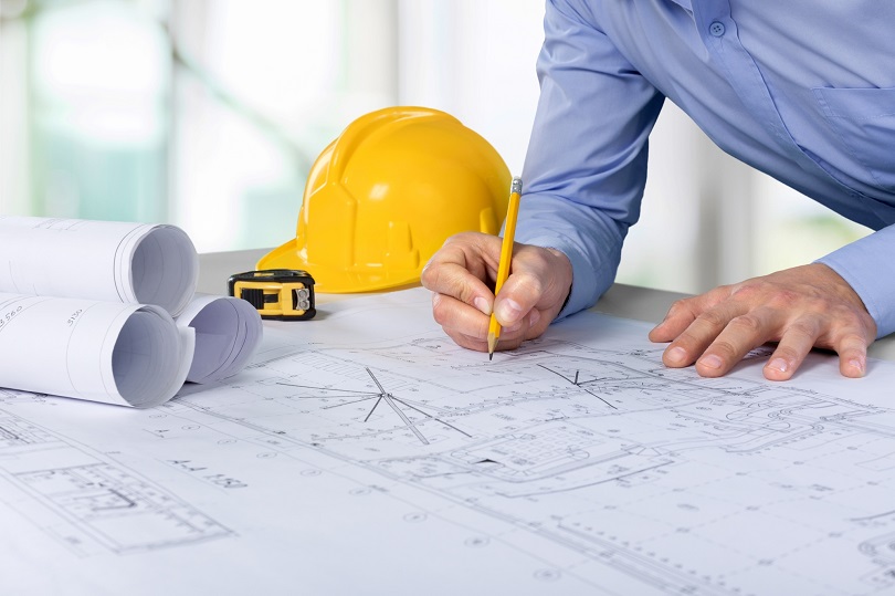 Foto ilustrativa em plano fechado com um homem escrevendo em uma planta de engenharia industrial que está sobre uma mesa. Ao lado, tambem há um capacete amarelo.