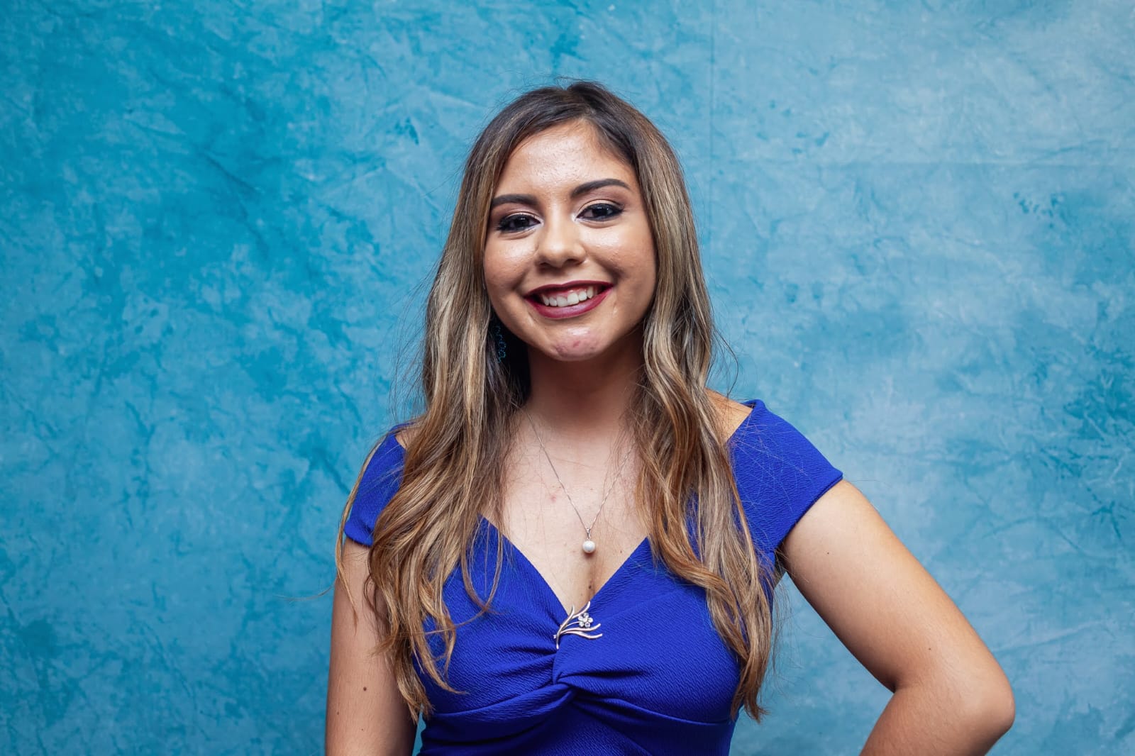 Foto de Amanda. Ela está sorrindo. Ela tem a pele clara e cabelos loiros, longos e ondulados. Está com maquiagem em tom dourado e batom de tonalidade marrom. Ela usa vestido azul e um colar dourado com uma pequena pedra clara. Ao fundo, há uma parede azul clara.