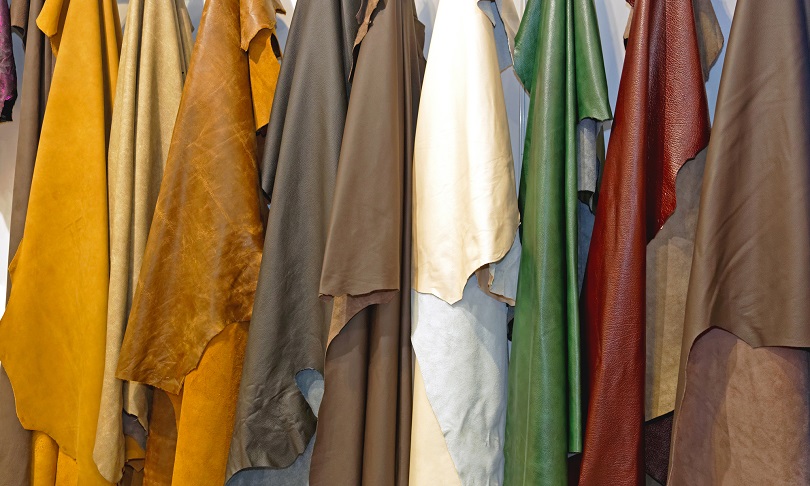 Foto ilustrativa em plano fechado sobre peças de couro de boi penduradas em um expositor. Os pedações de couro estão brutos e possuem diferentes cores: amarelo claro, amarelo escuro, marrom, branco, verde e vermelho. 