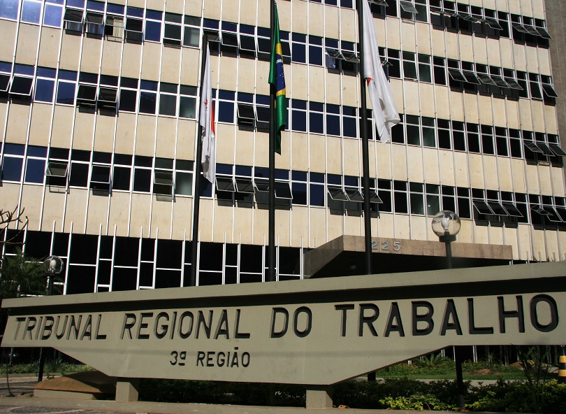 Parte do Prédio-Sede do TRT-3. A construção tem cor bege e, à frente, há três bandeiras hasteadas. É possível identificar apenas a do meio, que é a do Brasil, em cor verde, azul e amarelo. As demais têm a cor branca e não é possível identificá-las. Abaixo do prédio, há um grande letreiro construído em um painel de concreto: Tribunal Regional do Trabalho - 3ª Região.