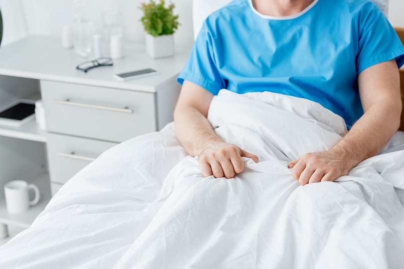 Foto ilustrativa, em plano fechado, de um homem que está de roupa cirúrgica azul, sentado em um leito de hospital. Ele está coberto nas pernas por uma coberta branca.