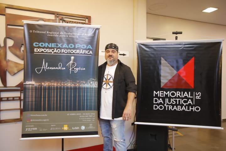 Foto do artista Alesxandro Rogério no meio de banners do Memorial da Justiça do Trabalho do RS e da mostra "Conexão Poa".