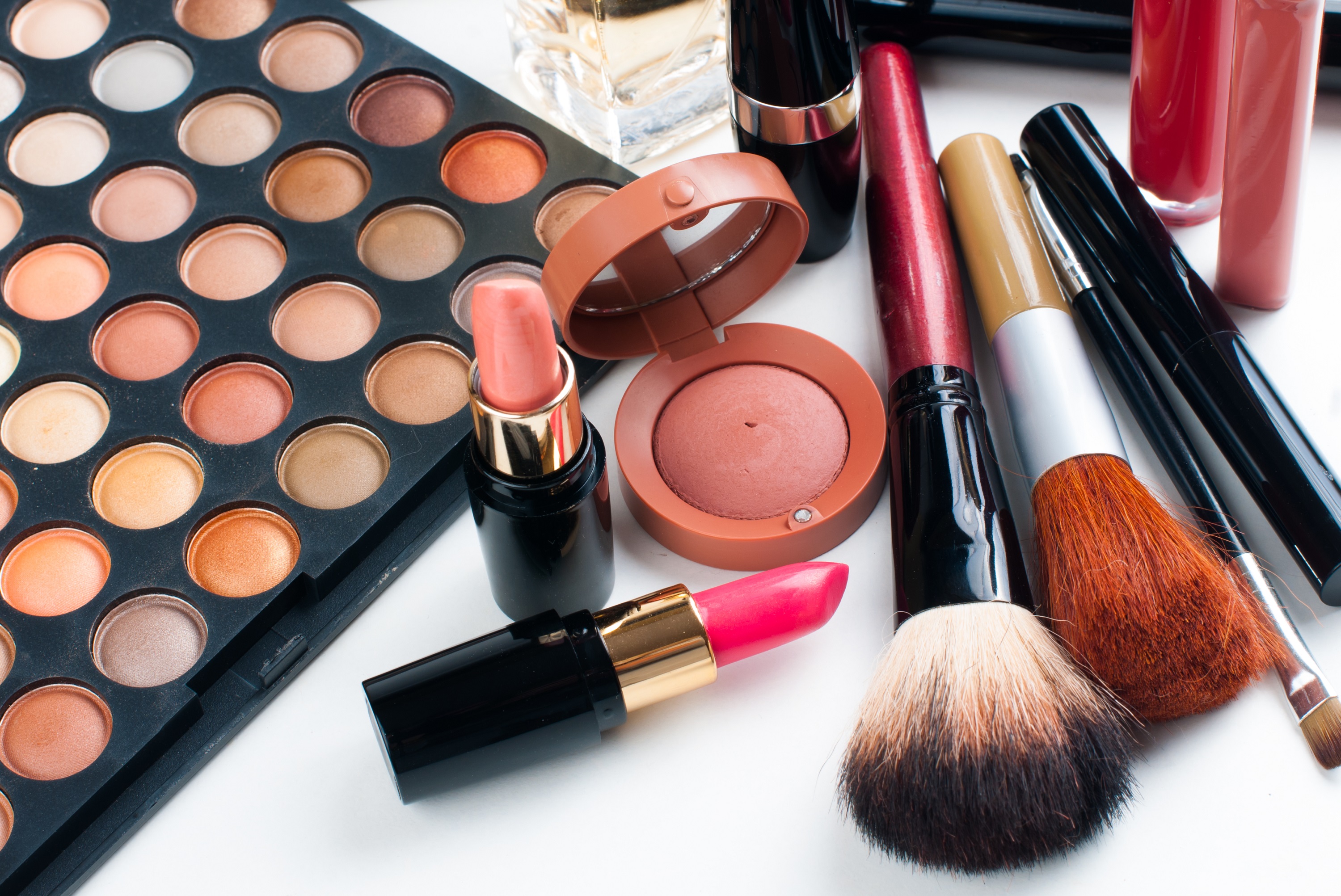 Estojo de maquiagens e batons em diversas cores sobre uma bancada branca. Há também um estojo de pó facial e pincéis de maquiagem, de tamanhos variados.