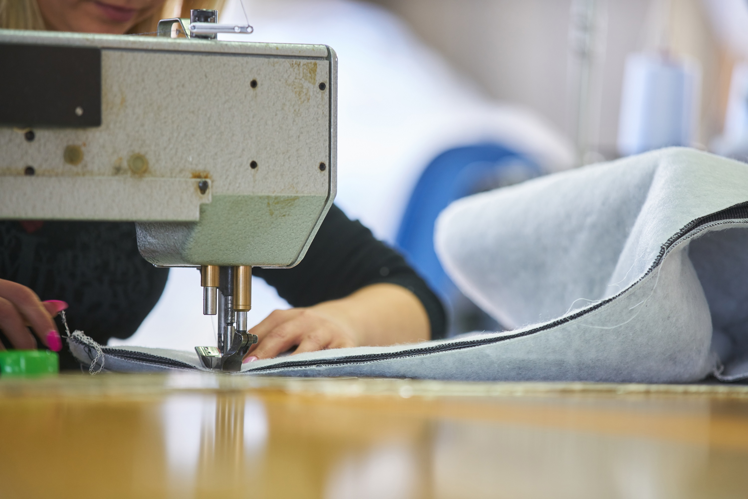 Costureira industrial costura forro, de cor cinza, em máquina industrial, de cor bege. Ela tem os cabelos loiros e usa esmalte cor de rosa nas mãos. O rosto dela está encoberto pela máquina.