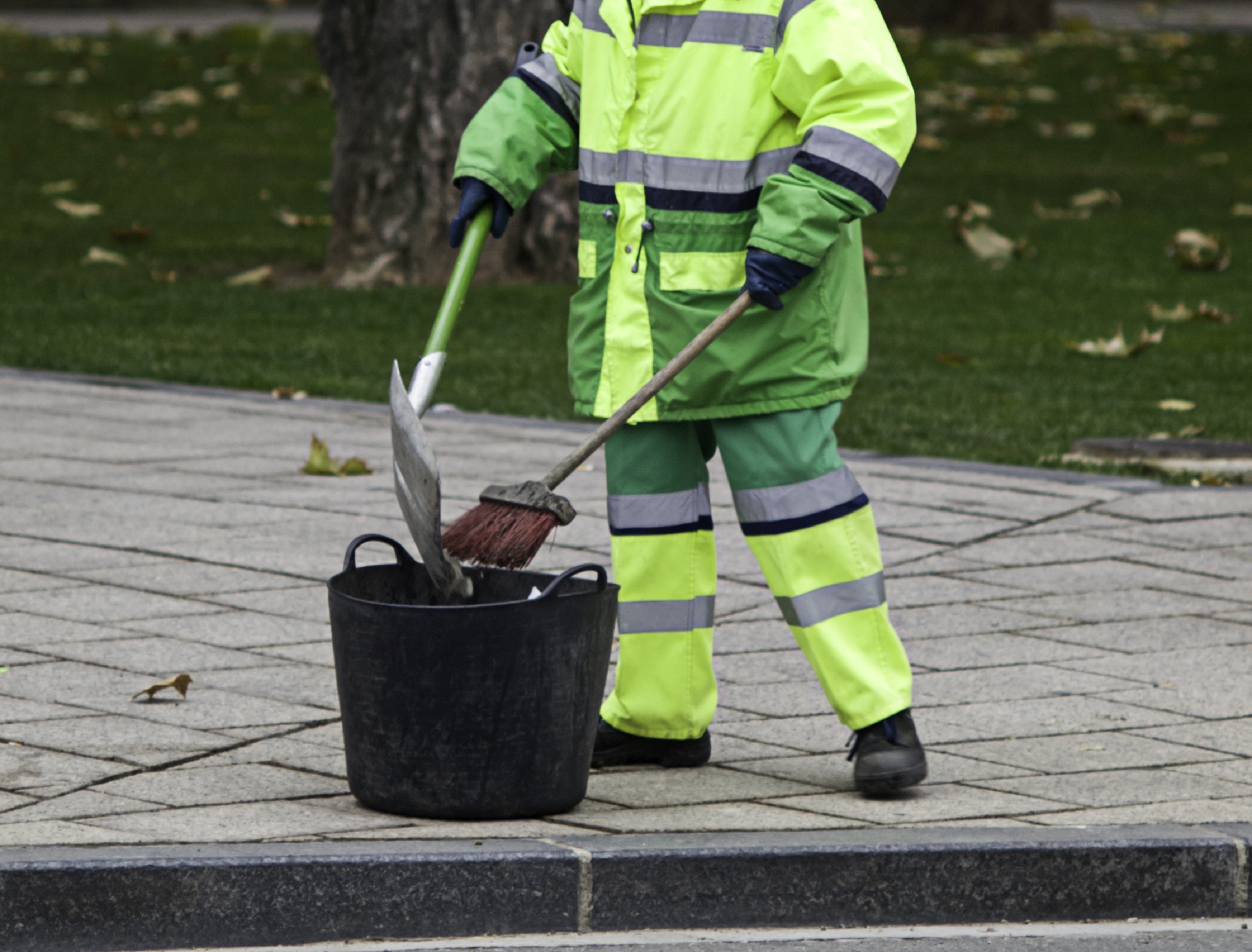 Homem com uniforme verde e cinza varrendo calçada. Não há imagem do rosto. Ele coloca o lixo em um recipiente preto.