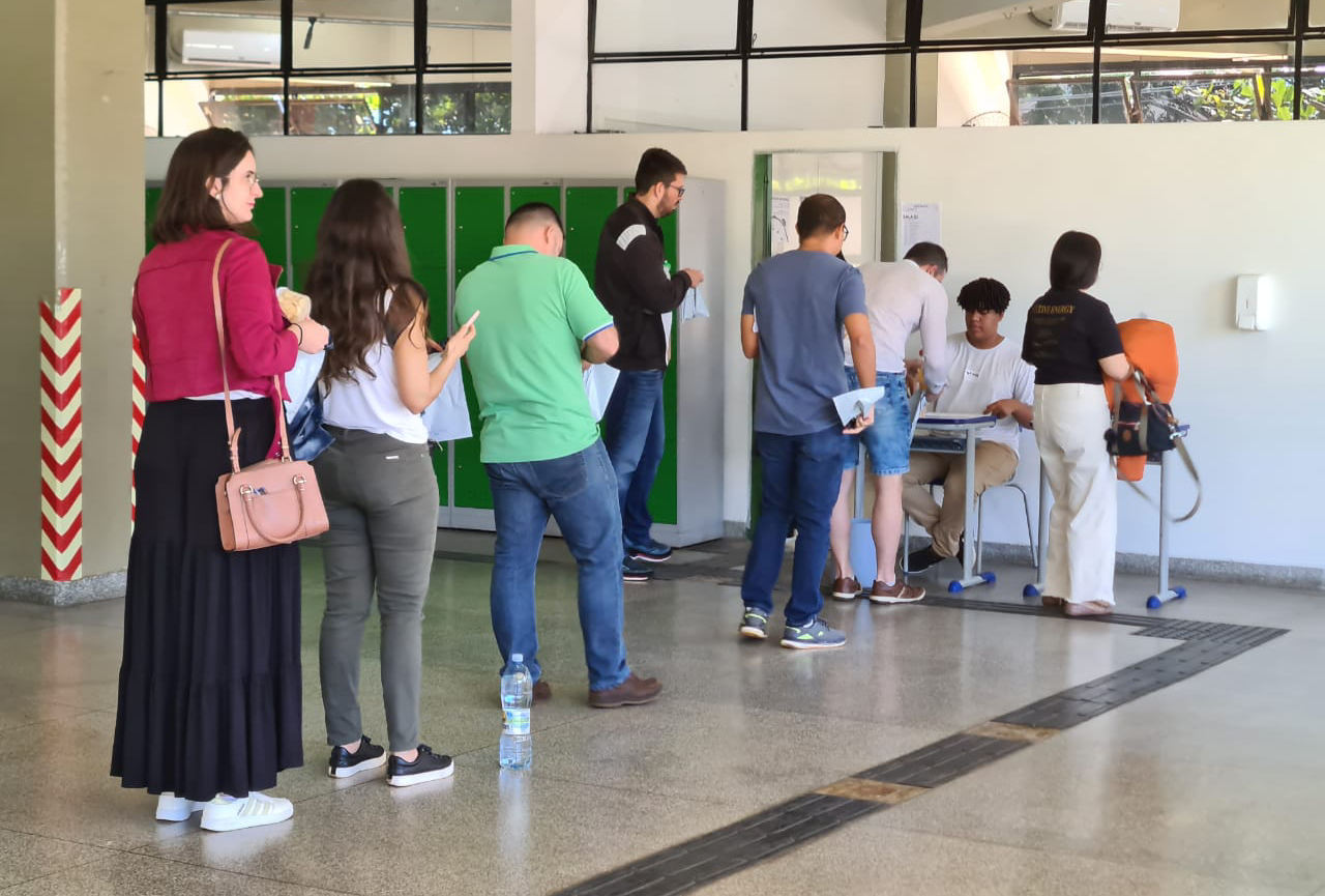 Fila com candidatas e e candidatos para entrar na sala de aplicação de prova. Homem sentado em carteira coletando as informações