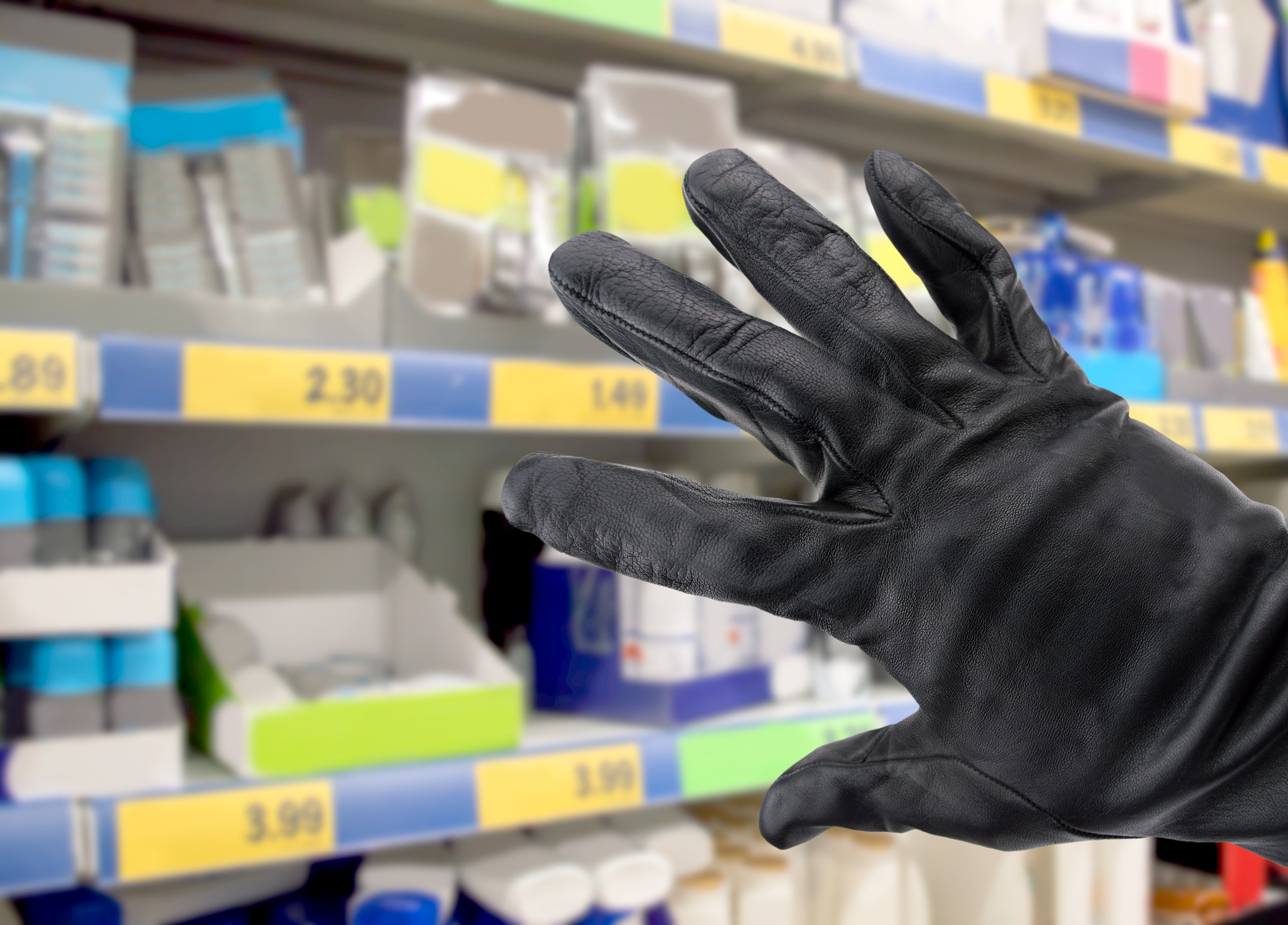 Mão com luva preta para impedir reconhecimento de digitais. Ao fundo, há gôndolas de supermercado. 
