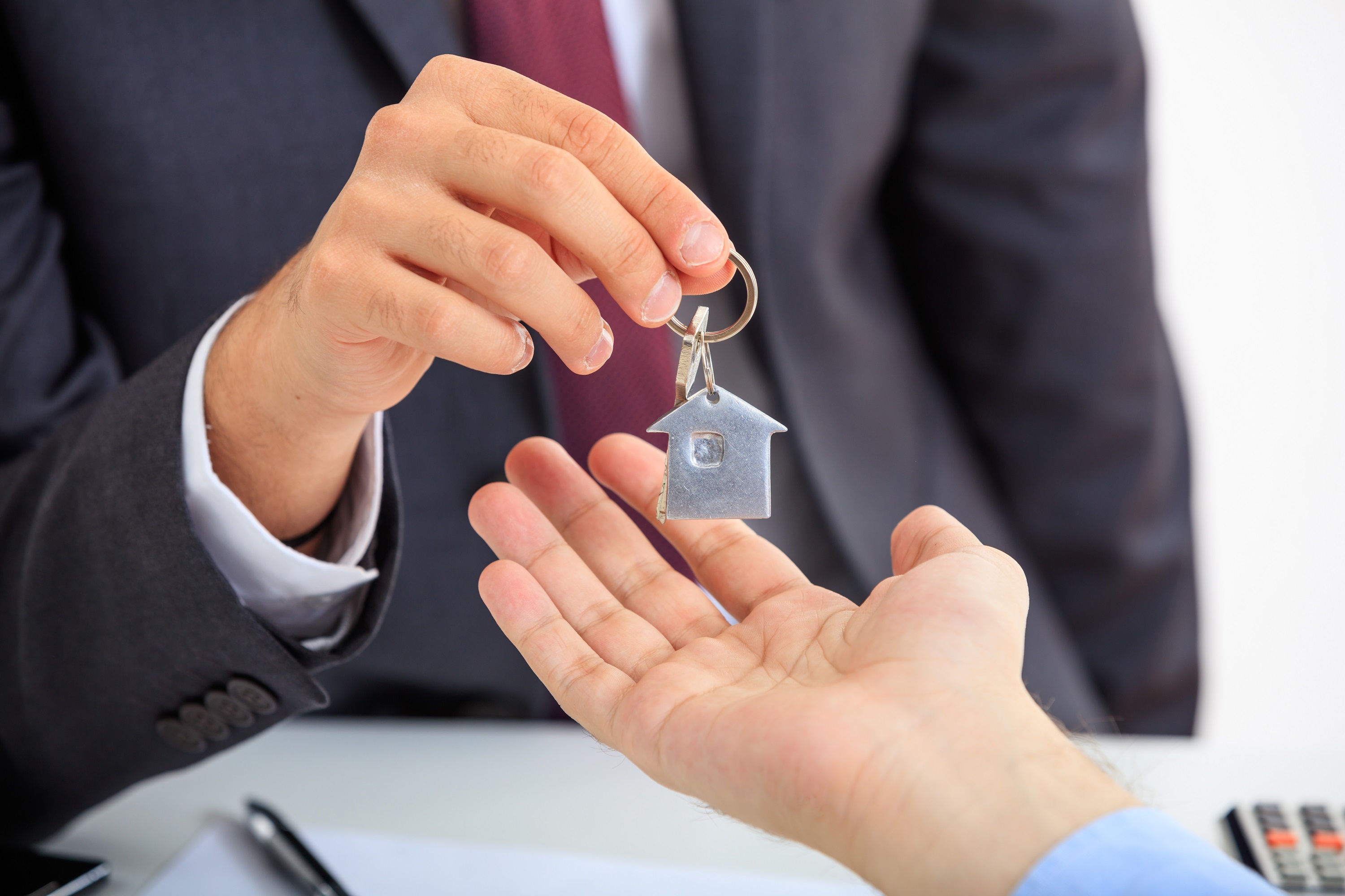 Homem entrega chave, com chaveiro em formato de casa, a outro homem. Aparecem apenas mãos. O homem que entrega a chave usa terno e gravata. 