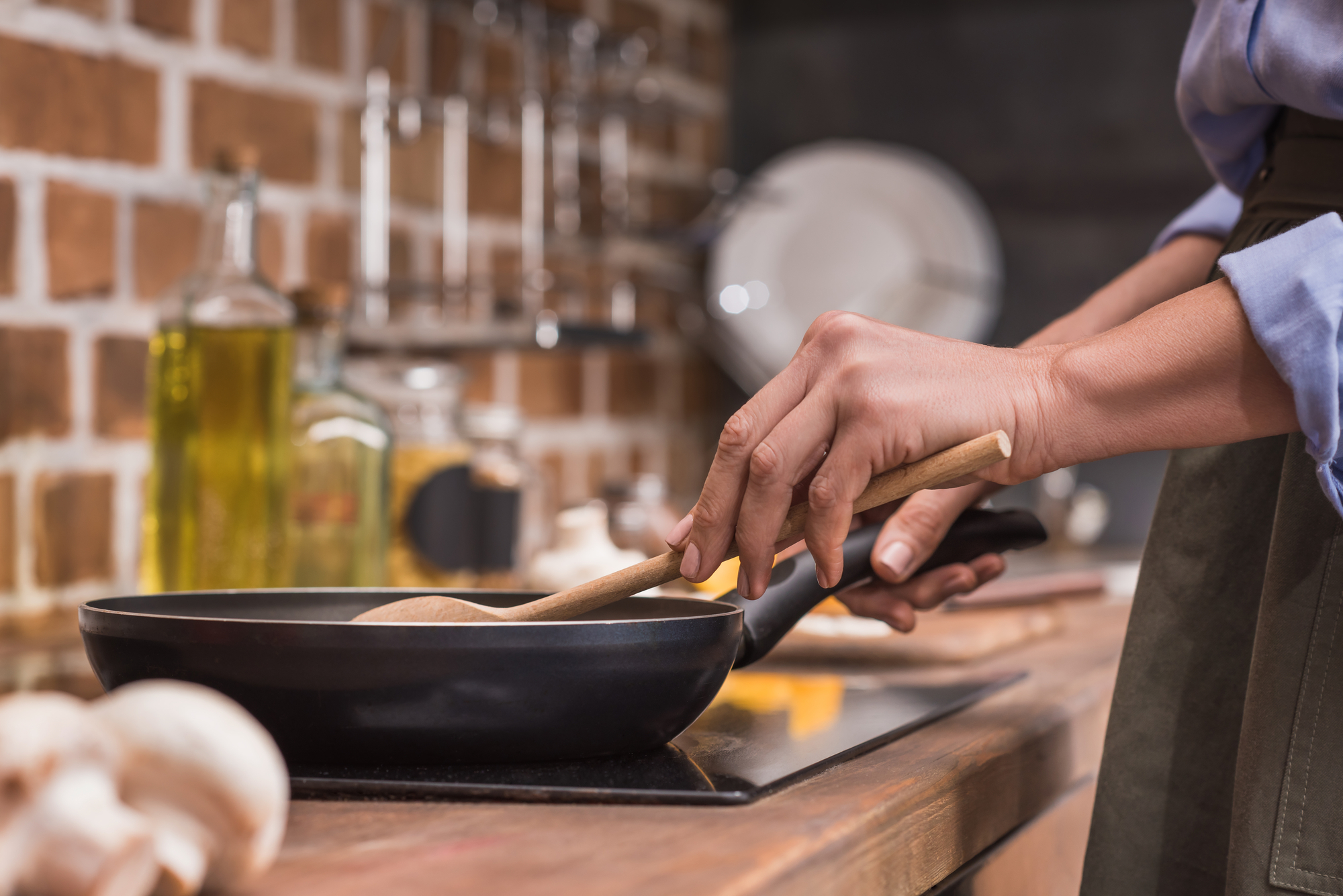 Foto ilustrativa das mãos de uma cozinheira com uma colher e uma frigideira. No cenário aparecem a bancada e a parede de uma cozinha.