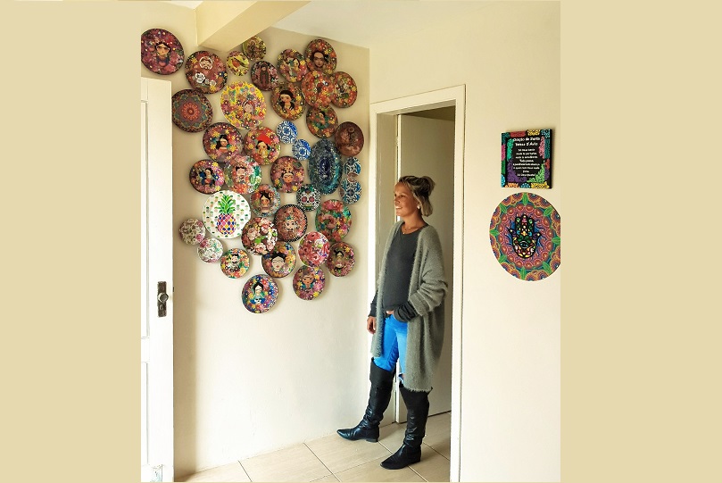 Lauren Robe em frente à parede com os pratos coloridos pendurados. As obras foram criadas por ela. Ela é uma mulher alta, pele morena e cabelos loiros, que estão presos. Usa calça jeans, botas pretas de cano longo, blusa e casaco cinza.