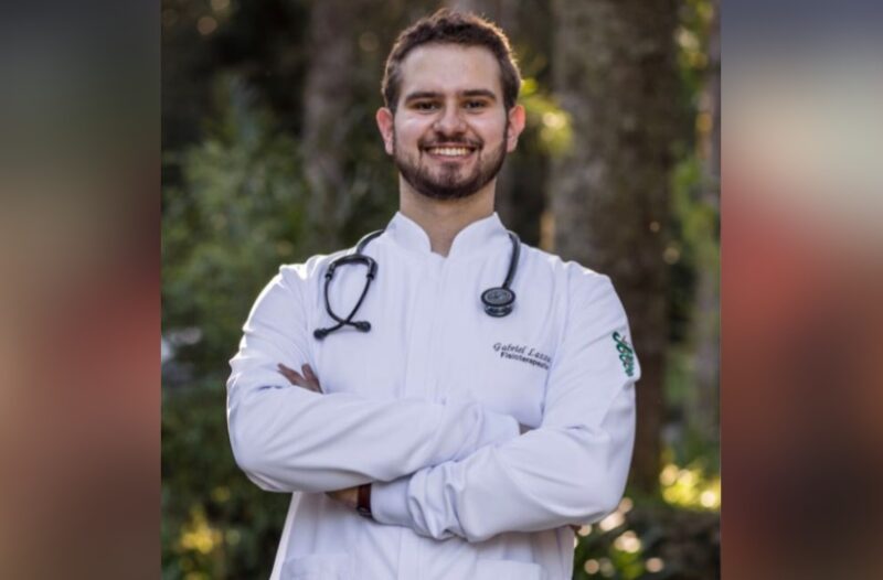 Foto de Gabriel Lazzari. Ele é branco, tem cabelo e barba curtos e castanhos, e usa um jaleco branco de profissional da saúde com um estetoscópio pendurando no pescoço,. Ele está de braços cruzados sorrindo para a fot.