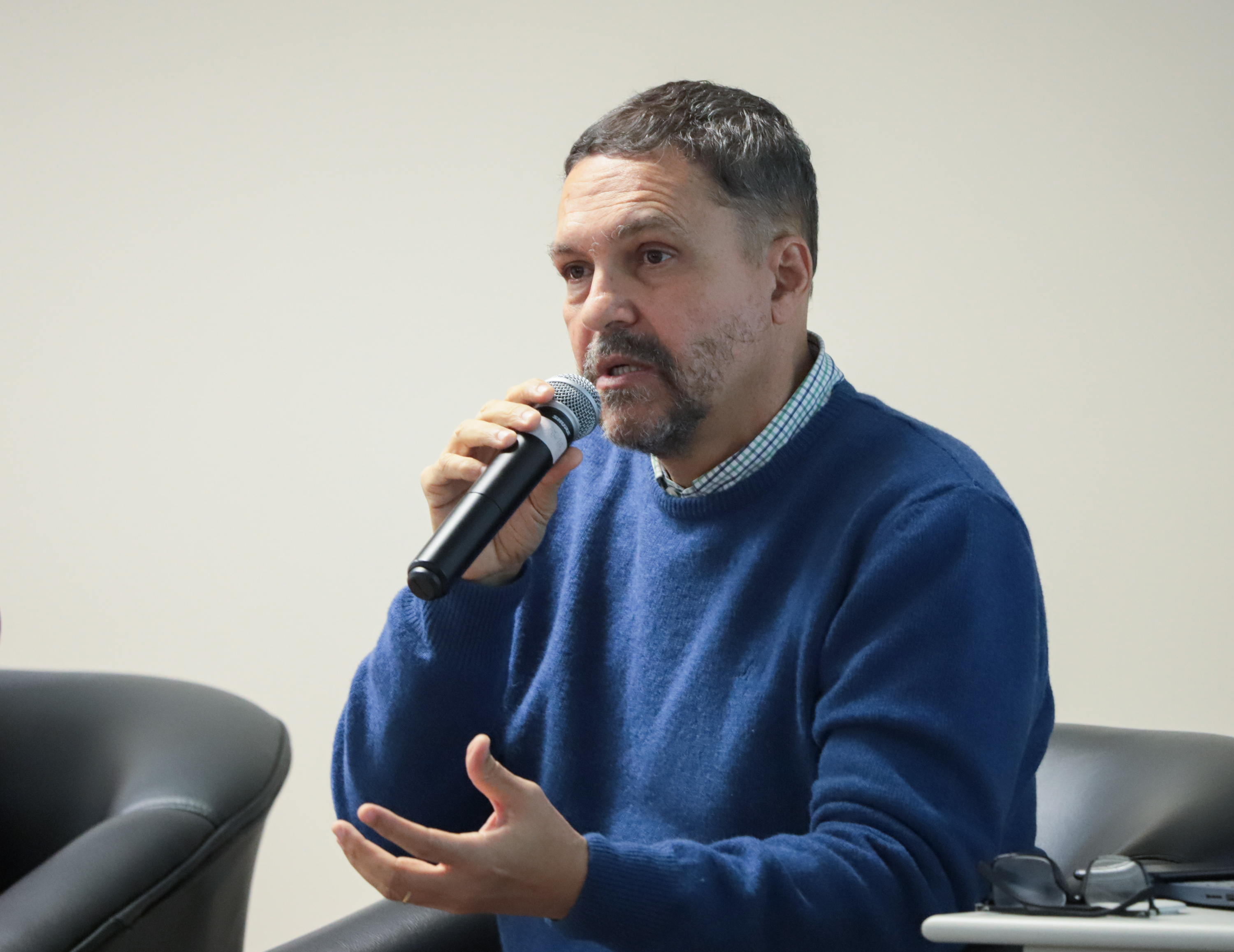 Paulo Scott é pardo, usa cabelo preto curto e barba curta. Está usando um blusão azul marinho e fala segurando um microfone.