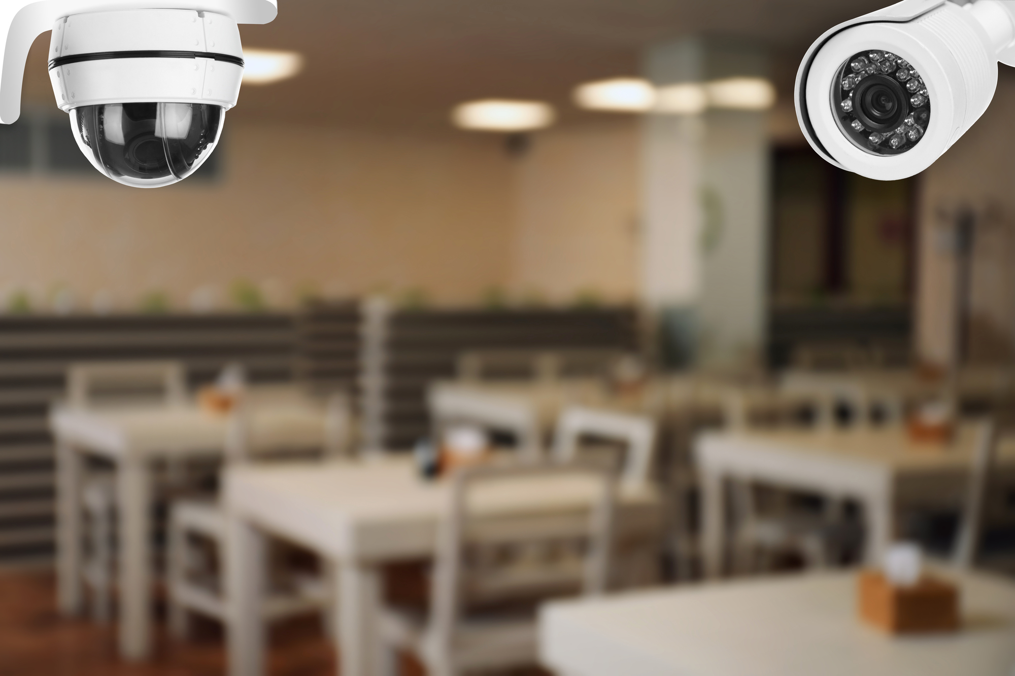 Câmeras de vigilância em um refeitório. As câmeras são brancas e o refeitório em tons claros de bege e branco.
