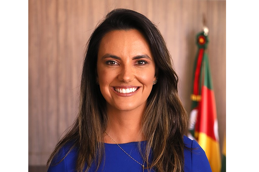 Retrato da juíza Carolina Gralha. Ela é branca, tem cabelo liso escuro e olhos escuros. Está sorrindo para a foto. Usa uma blusa azul e um colar dourado. Ao fundo aparece a bandeira do RS.