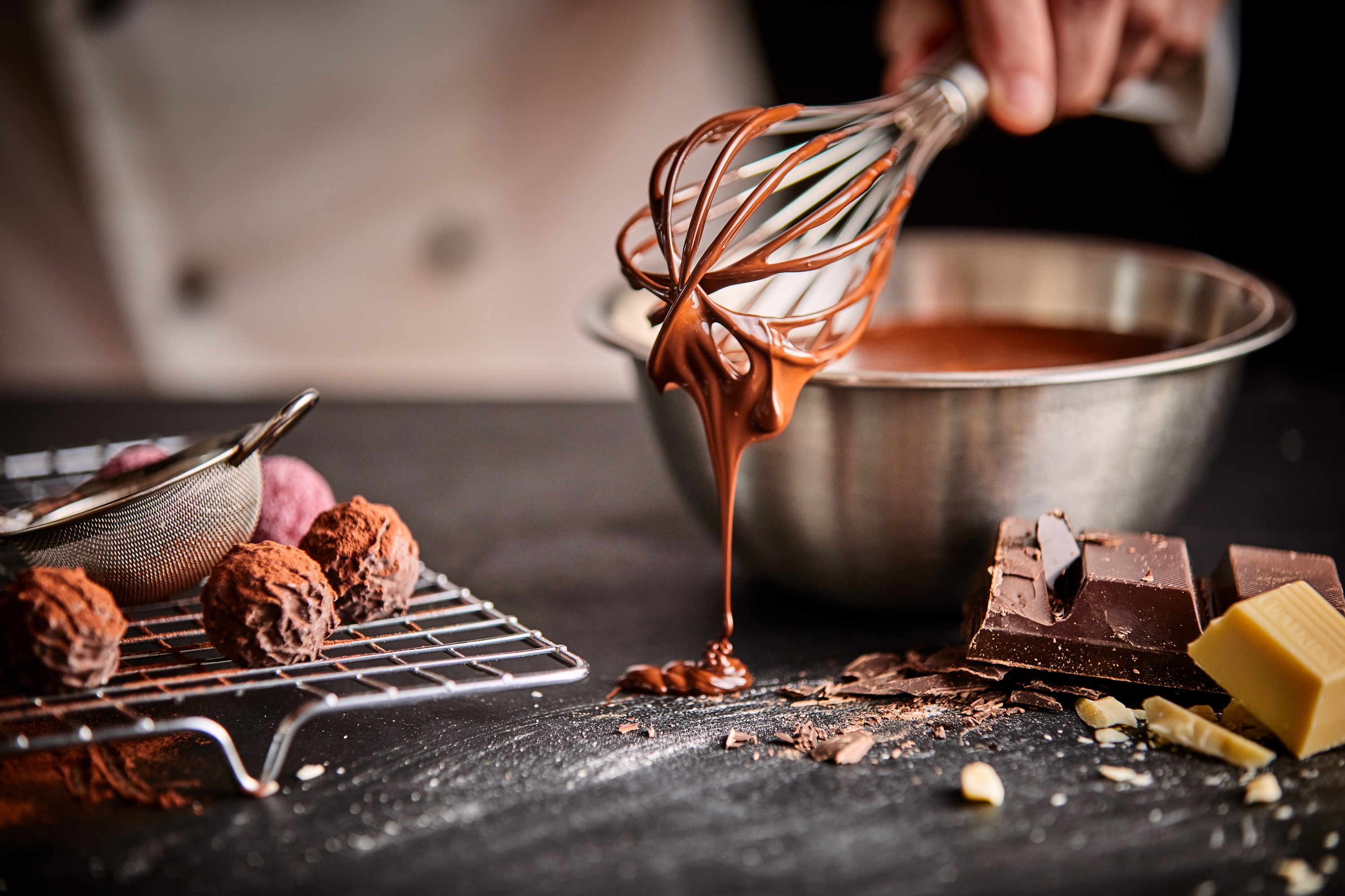 Confeiteira prepara chocolates. Apenas a mão segurando um batedor (fouet) é mostrada. Há uma tigela com chocolate e outros doces sobre a mesa.