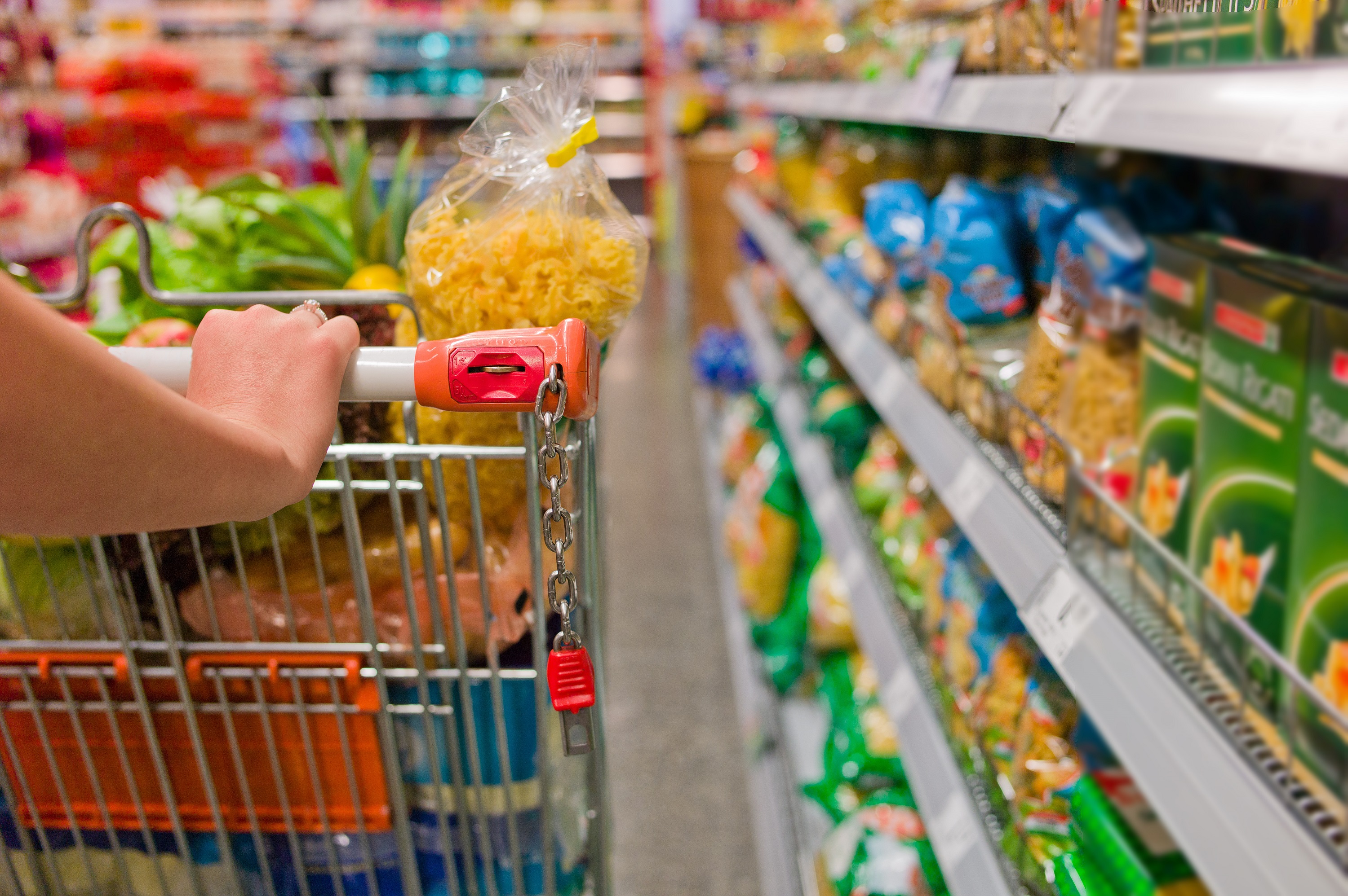 Mulher com carrinho de compras no supermercado. Não há a imagem do rosto. Ao lado do carrinho, há prateleiras com mercadorias