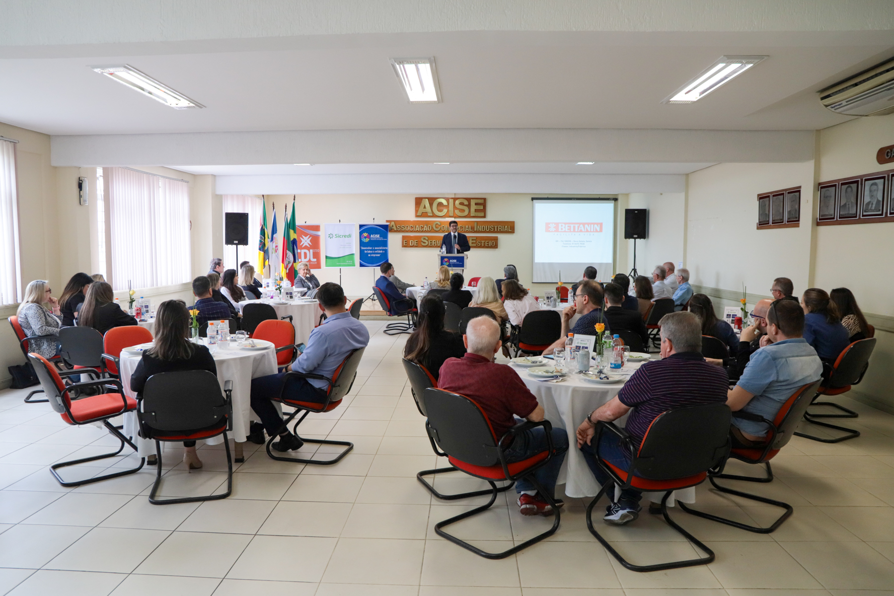 Foto em ângulo aberto do evento, mostrando público sentado (mesas redondas) e o desembargador falando, ao fundo