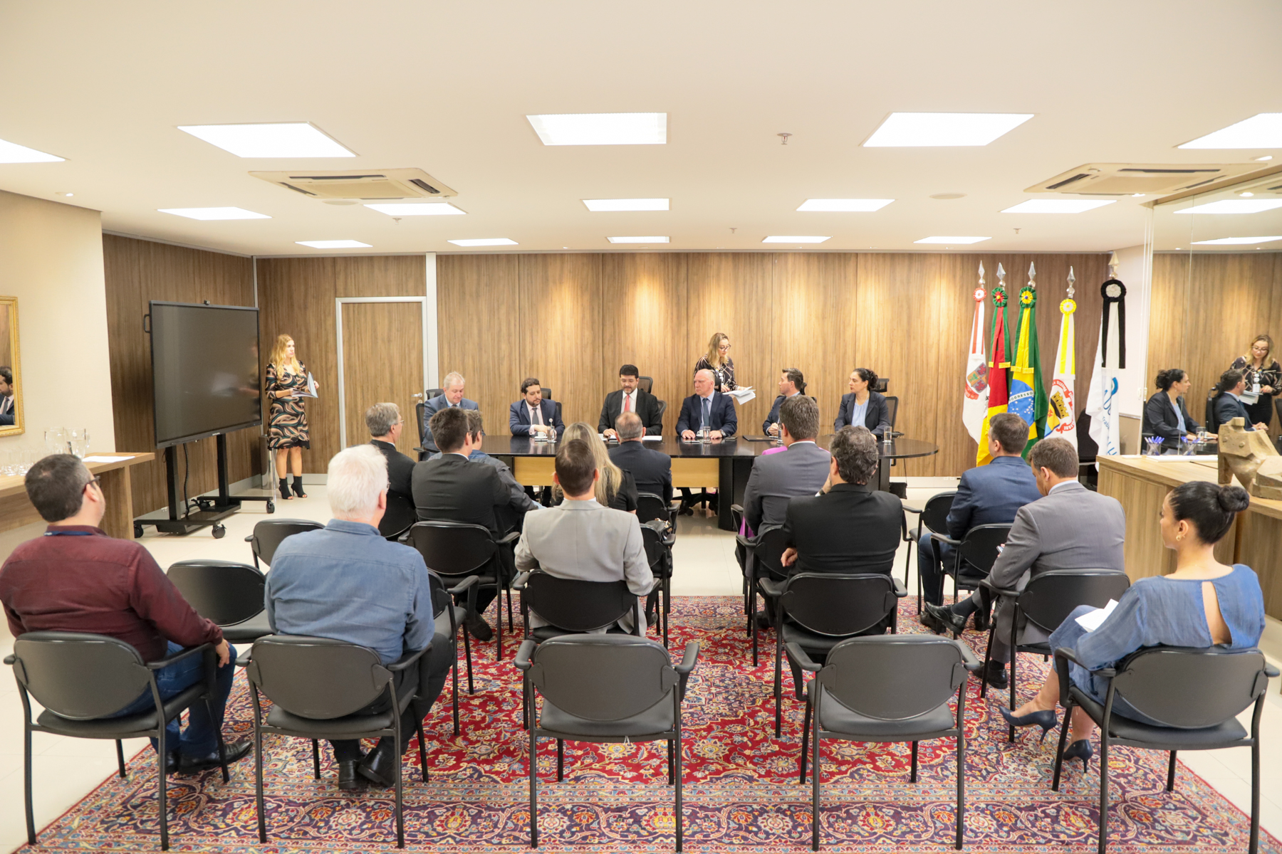 Foto em ângulo aberto, mostrando a mesa oficial e demais pessoas sentadas em cadeiras