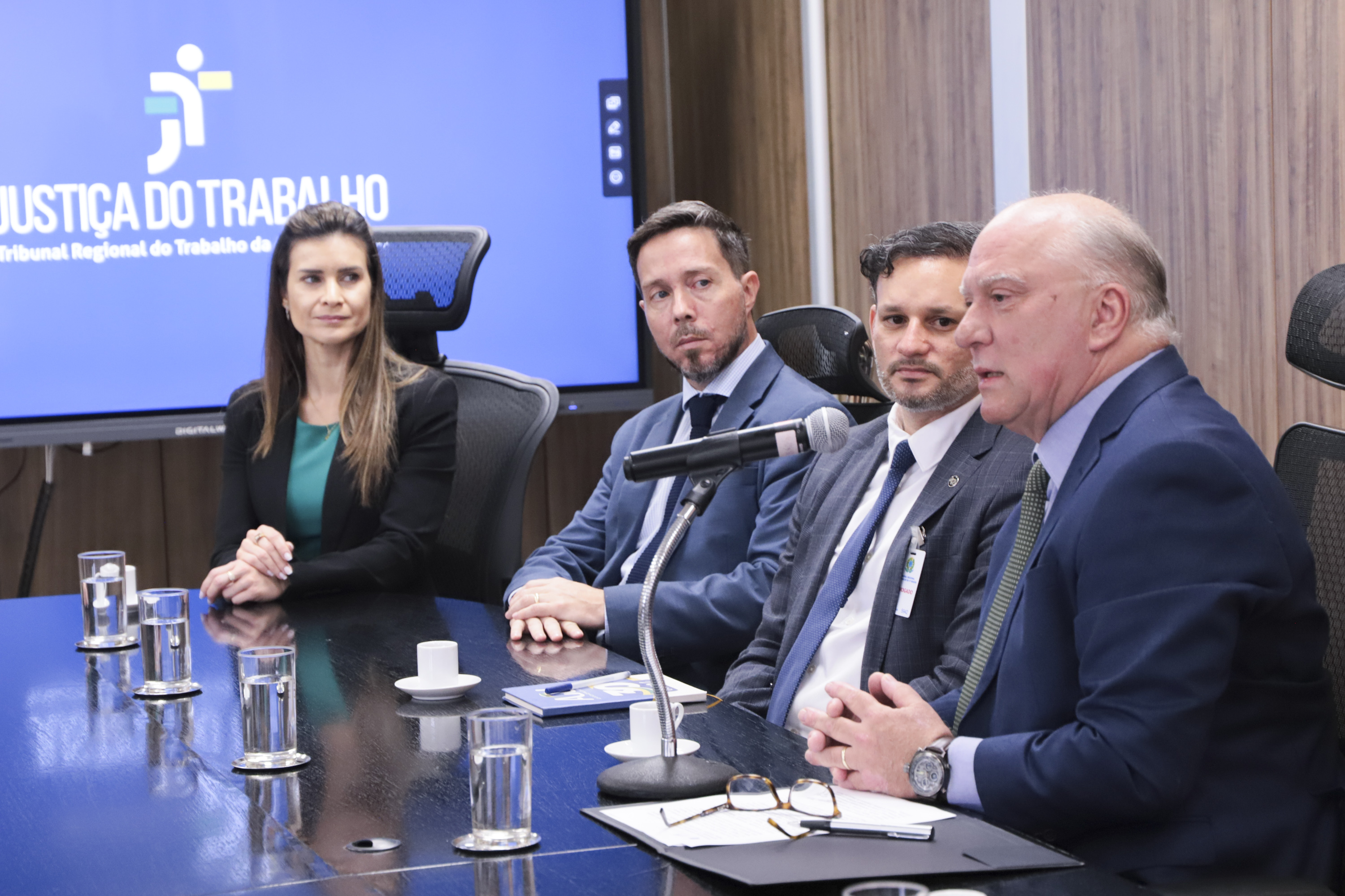 Martins Costa fala ao microfone em primeiro plano, à direita da foto. Ao fundo aparecem Melquizedeck, Trindade e Aline Lima, e uma tela com o logo da JT