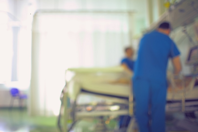 Imagem desfocada de dois homens, vestindo uniforme azul, próximos à cama hospitalar. Ao fundo, há bastante luminosidade.