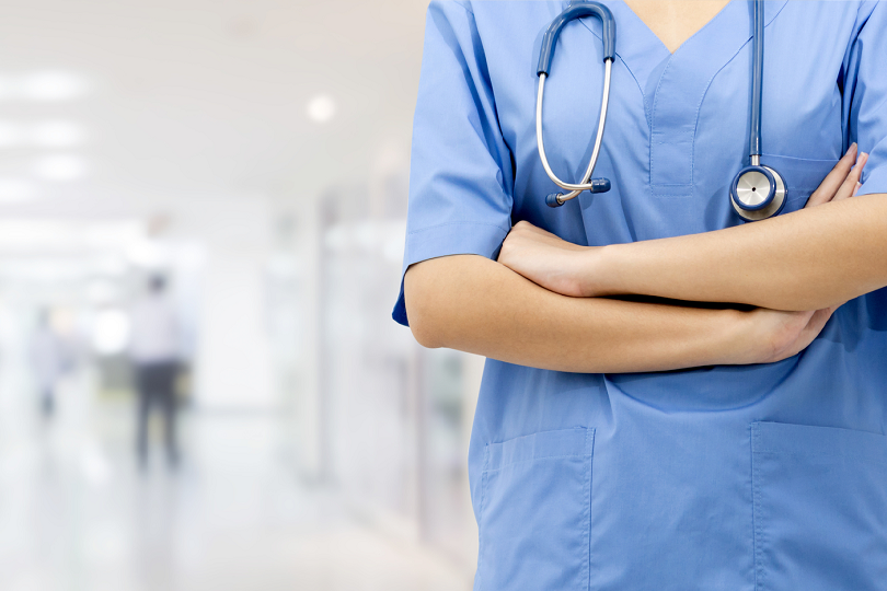 profissional de saúde em corredor de hospital, com os braços cruzados. Ela tem a pele clara e usa roupa toda azul. Tem um estetoscópio sobre os ombros.