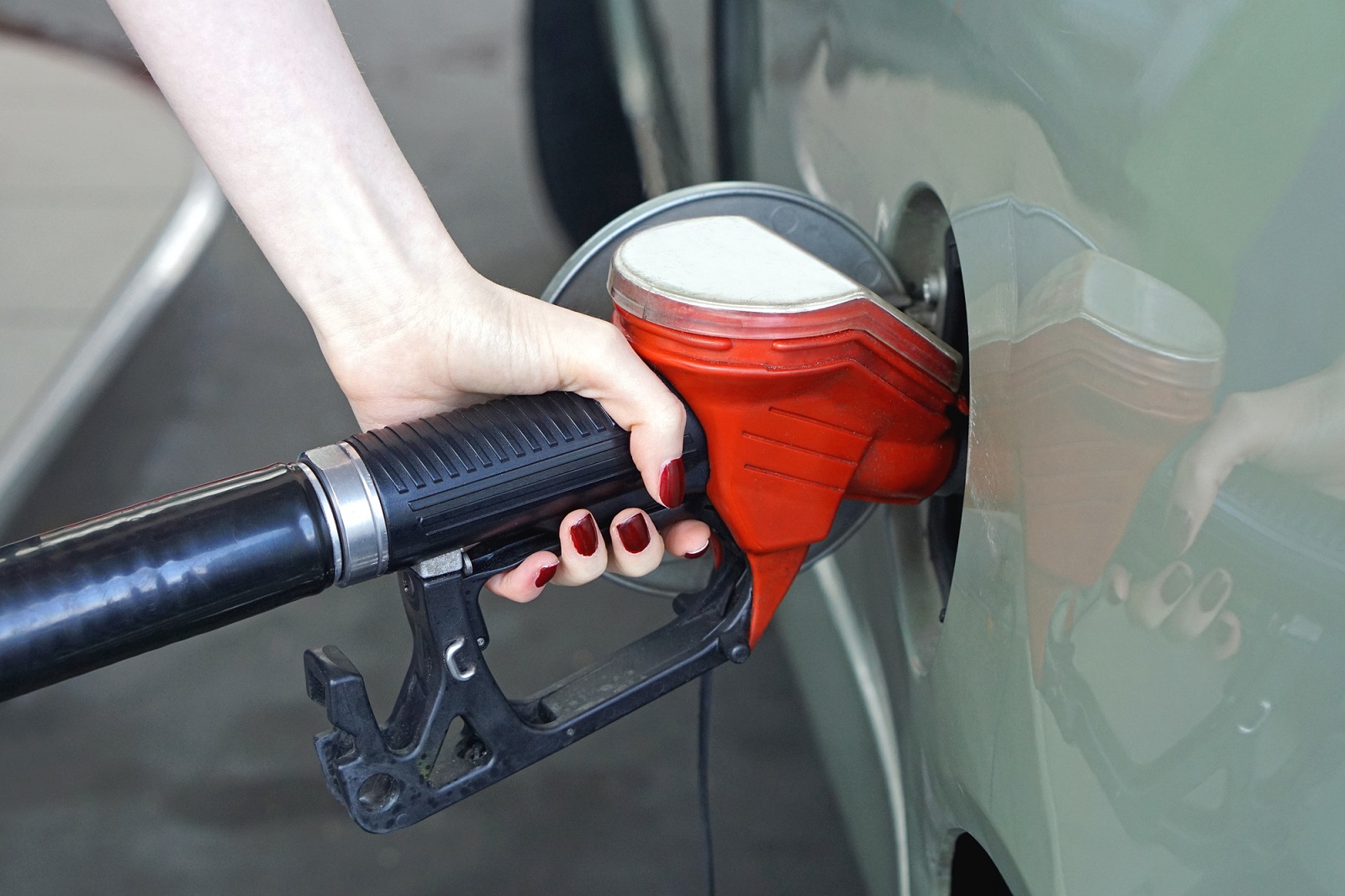 ãos femininas abastecem carro. O veículo tem a cor cinza. A pele da mulher é branca e as unhas estão pintadas de vermelho escuro. 