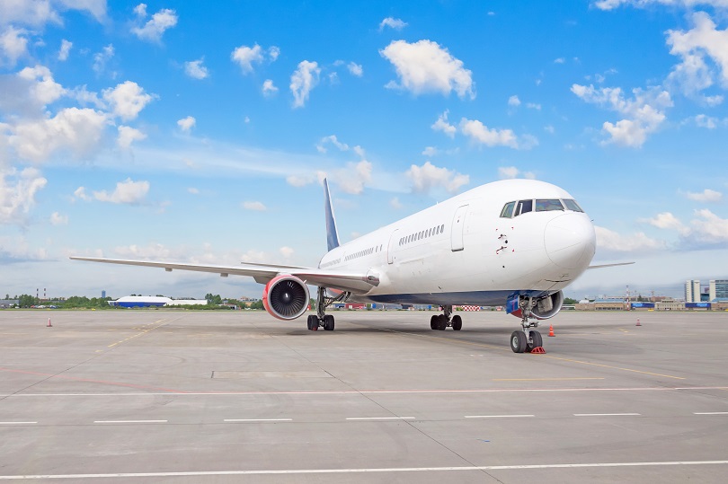 Avião, de cor branca, estacionado no aeroporto. Ao fundo, há o horizonte. 