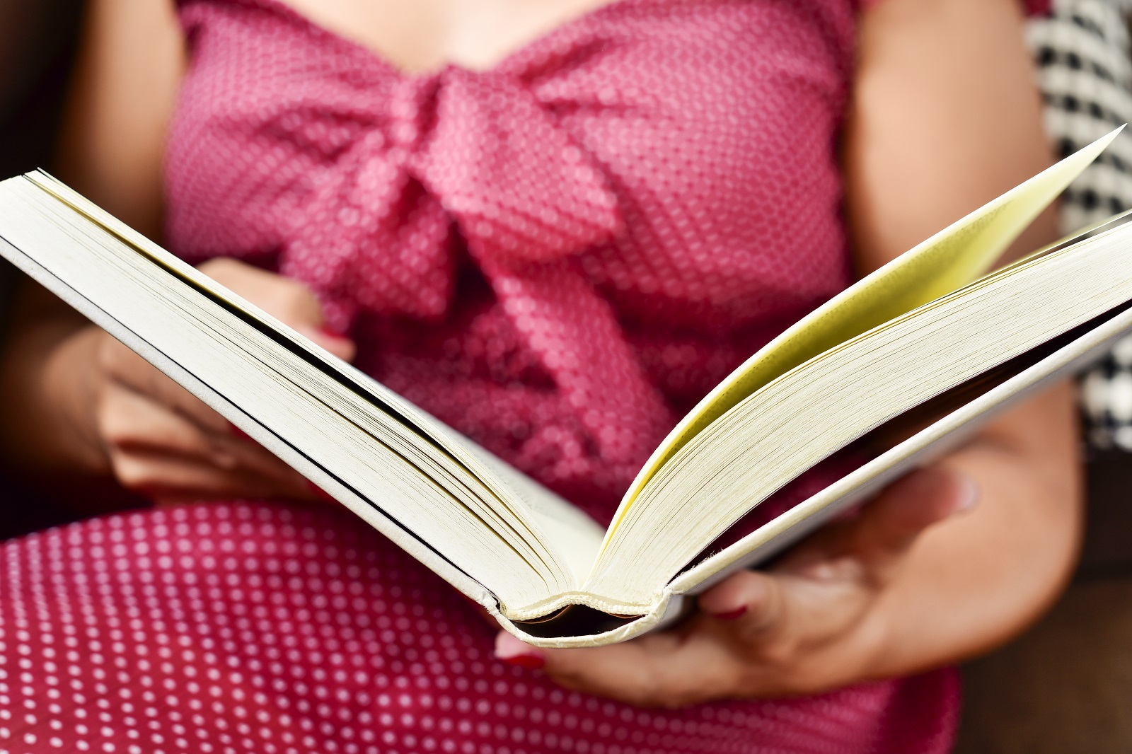 Foto em foco de livro aberto, uma mulher sentada vestindo um vestido vermelho com bolinhas brancas segura o livro, a mulher está levemente desfocada.