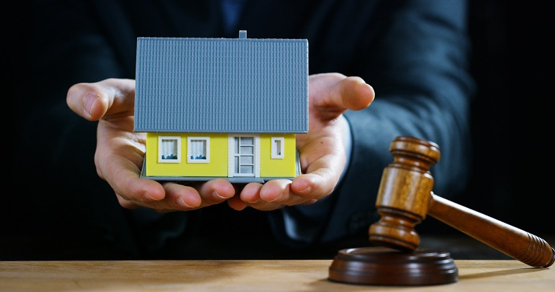 foto ilustrativa, em plano fechado, das mãos de um homem que segura a maquete de uma casa. Ao lado da maquete, está posicionado um martelo de leilão.