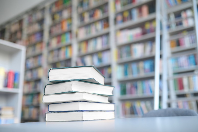 Foto ilustrativa de livros sobre uma mesa. Ao fundo, há estantes com livros.