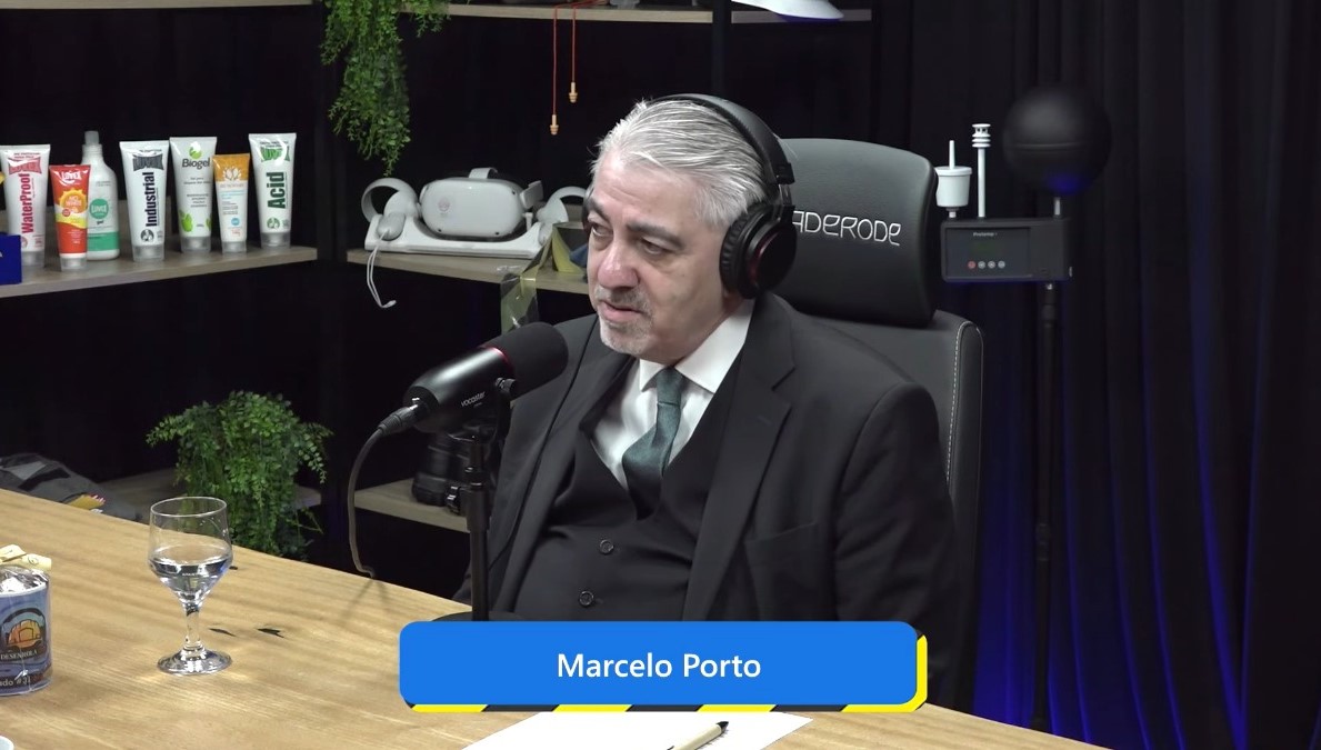 captura de tela do juiz Marcelo dando a entrevista no estúdio do podcast. Ele é um home de meia idade, cabelos e cavanhaque grisalhos, pele branca e olhos castanhos. Veste um terno escuro, colete da mesma cor,  camisa branca e gravata cinza.