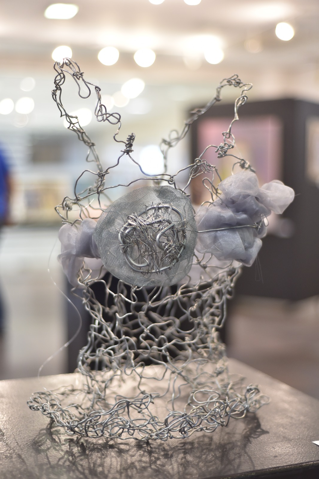 Foto em plano fechado de uma escultura em forma de corselete, feita com fio metálico prateado e materiais brilhantes