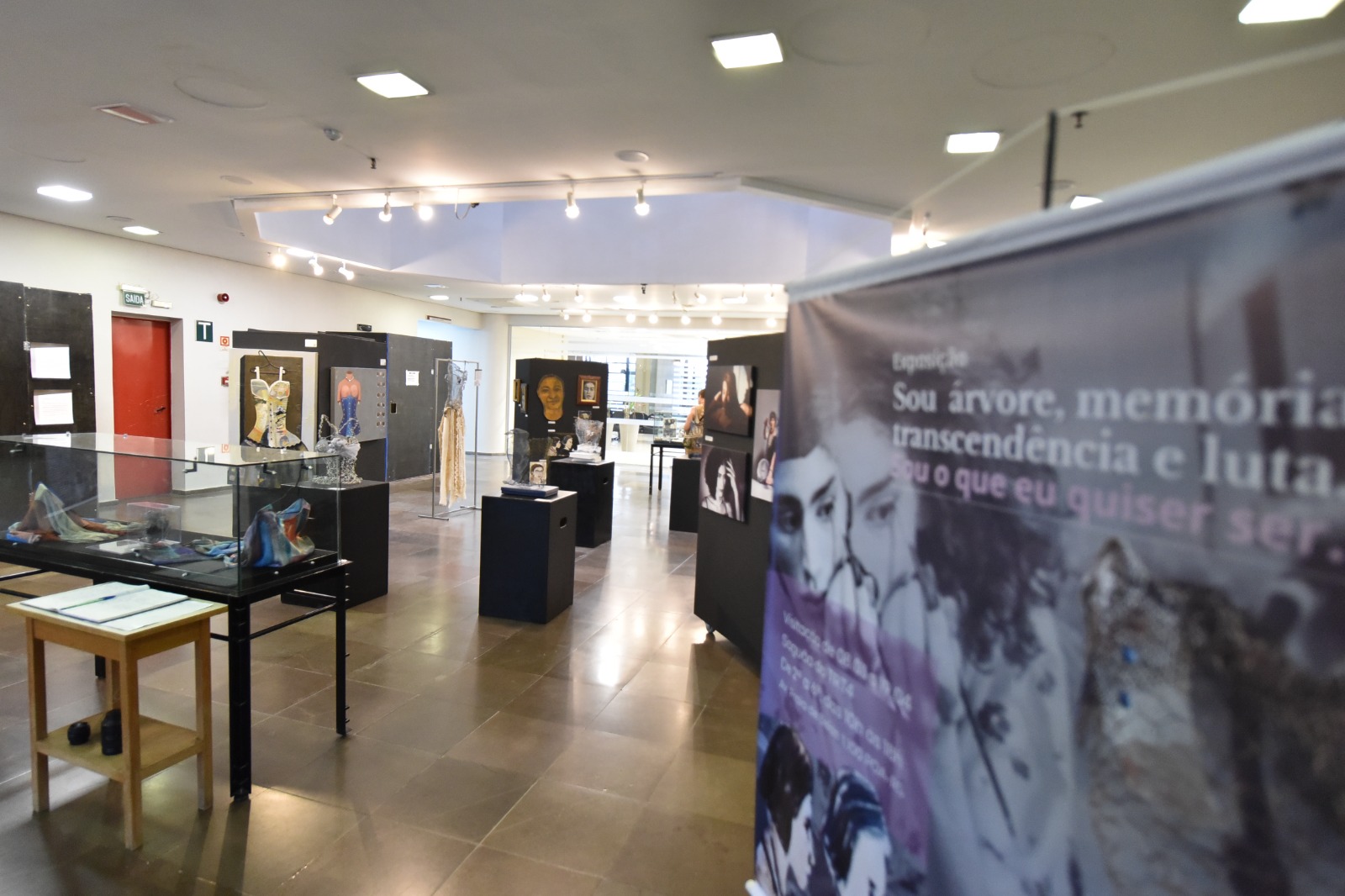 Foto em plano aberto do saguão, exibindo todas obras expostas e, em primeiro plano, o banner de divulgação, no qual consta o nome da exposição e fotos das obras