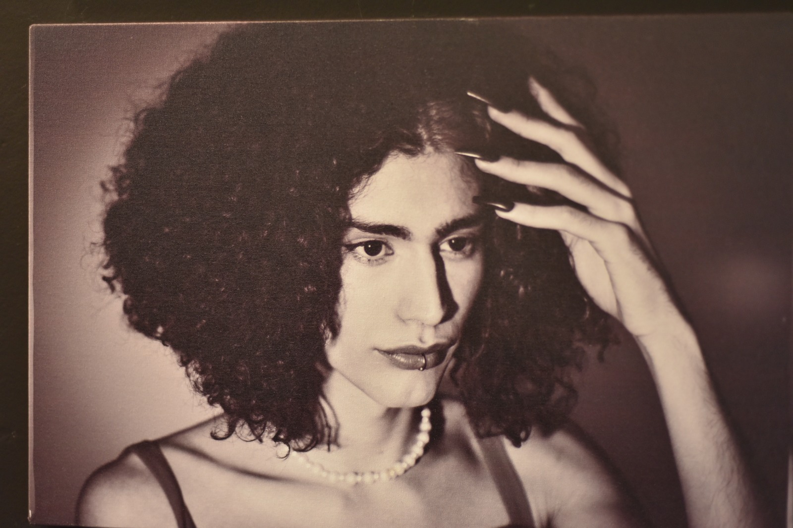 Foto de uma foto em preto e branco e plano fechado do rosto de uma mulher trans. Ela tem pele branca e cabelos encaracolados e volumosos, tocados pela sua mão esquerda que tenha unhas longas e pintadas. Usa um piercing de argola no centro do lábio inferior.