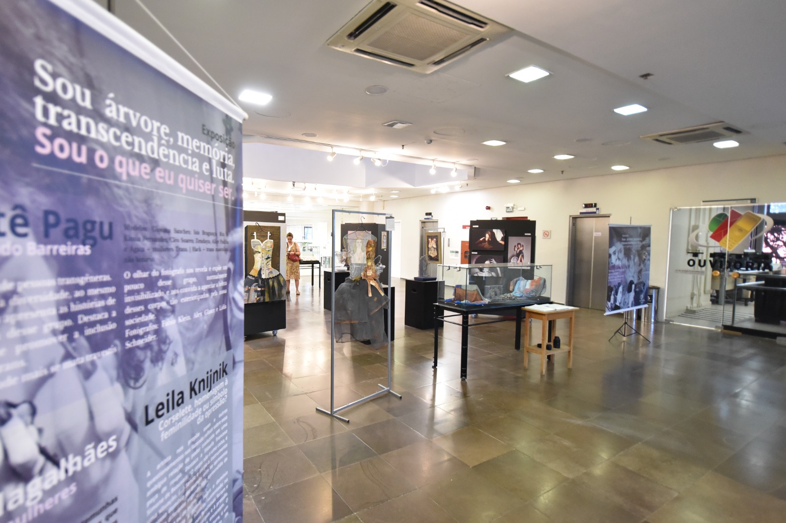 Foto em plano aberto do saguão, exibindo todas obras expostas e, em primeiro plano, o banner de divulgação, no qual consta o nome da exposição e fotos das obras
