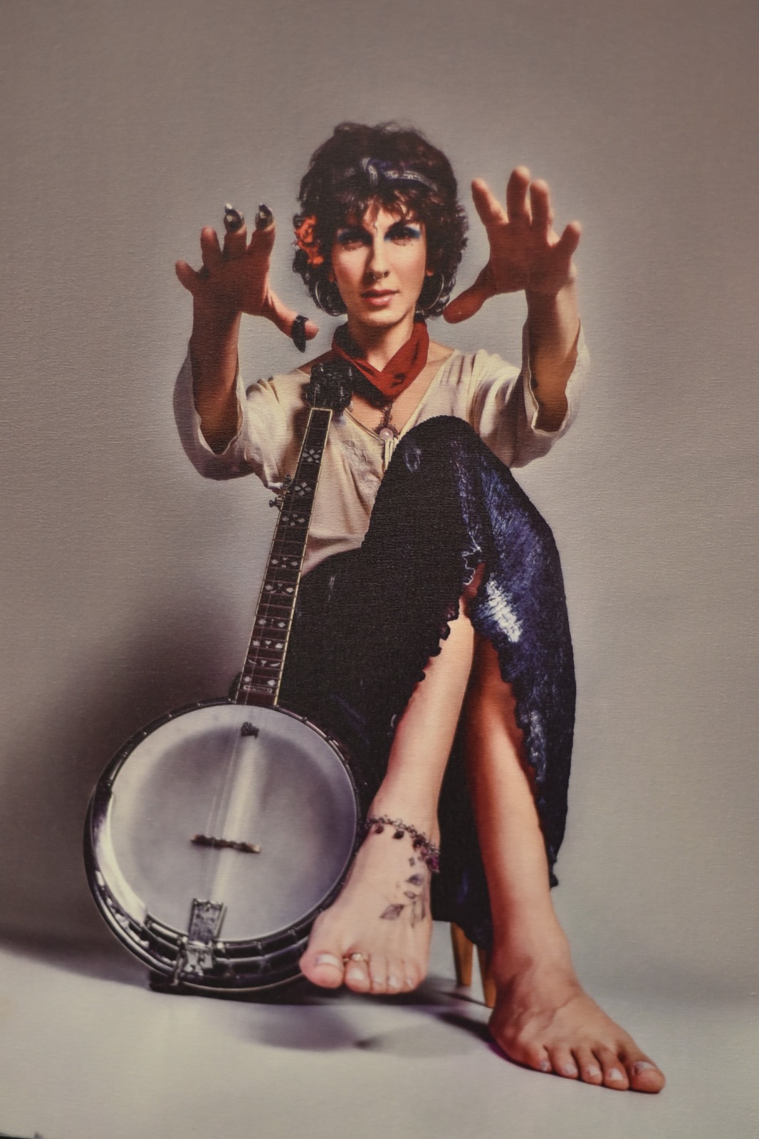 Foto de uma foto colorida de um/a músico/a sentado, olhando para a câmera, com um banjo americano encostado em seu corpo e suas mãos esticadas para frente, na direita vemos as dedeiras de metal usadas para dedilhar o instrumento. Os pés decalços aparecem em primeiro plano; no esquerdo, há uma tornozeleira, uma tatuagem de um ramo no peito e um anel em um dedo. tem a pele branca, cabelo castanho, sombra azul nos olhos, usa uma blusa clara, saia azul longa e tem as pernas cruzada, semi-flexionadas